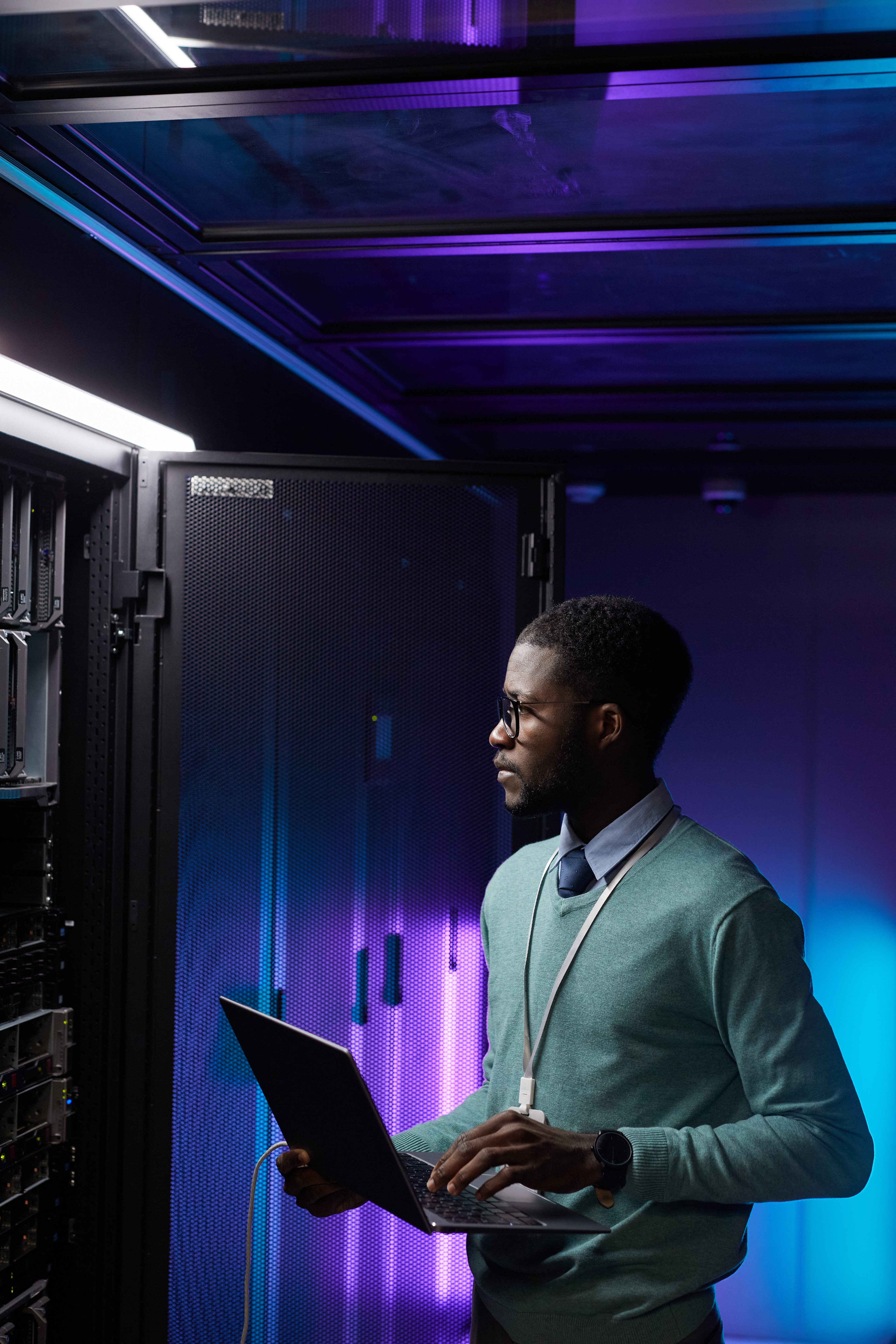 Vertical portrait of young African American data engineer holding laptop while working with supercomputer in server room lit by blue light, copy space